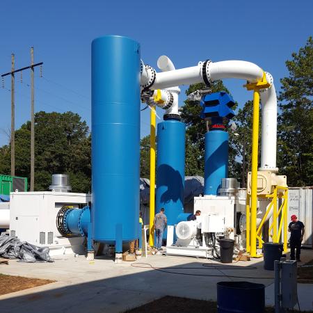 Outdoor industrial assembly of tall blue vacuum tanks and white piping with two workers standing nearby