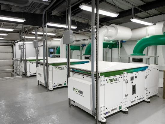 Row of white industrial dust collection units labeled 'inovair' with green ducting overhead in an indoor facility