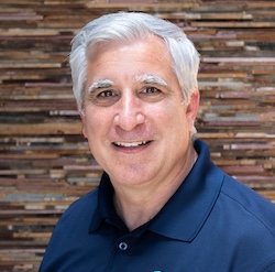 Headshot of a man in a navy polo shirt, smiling against a wood-paneled background