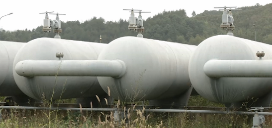 Three large white horizontal storage tanks with valve assemblies on top, photographed outdoors with trees in the background