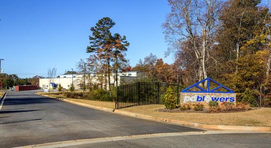 Entrance sign reading 'pd blowers' beside a driveway with landscaping, trees, and a building in the background